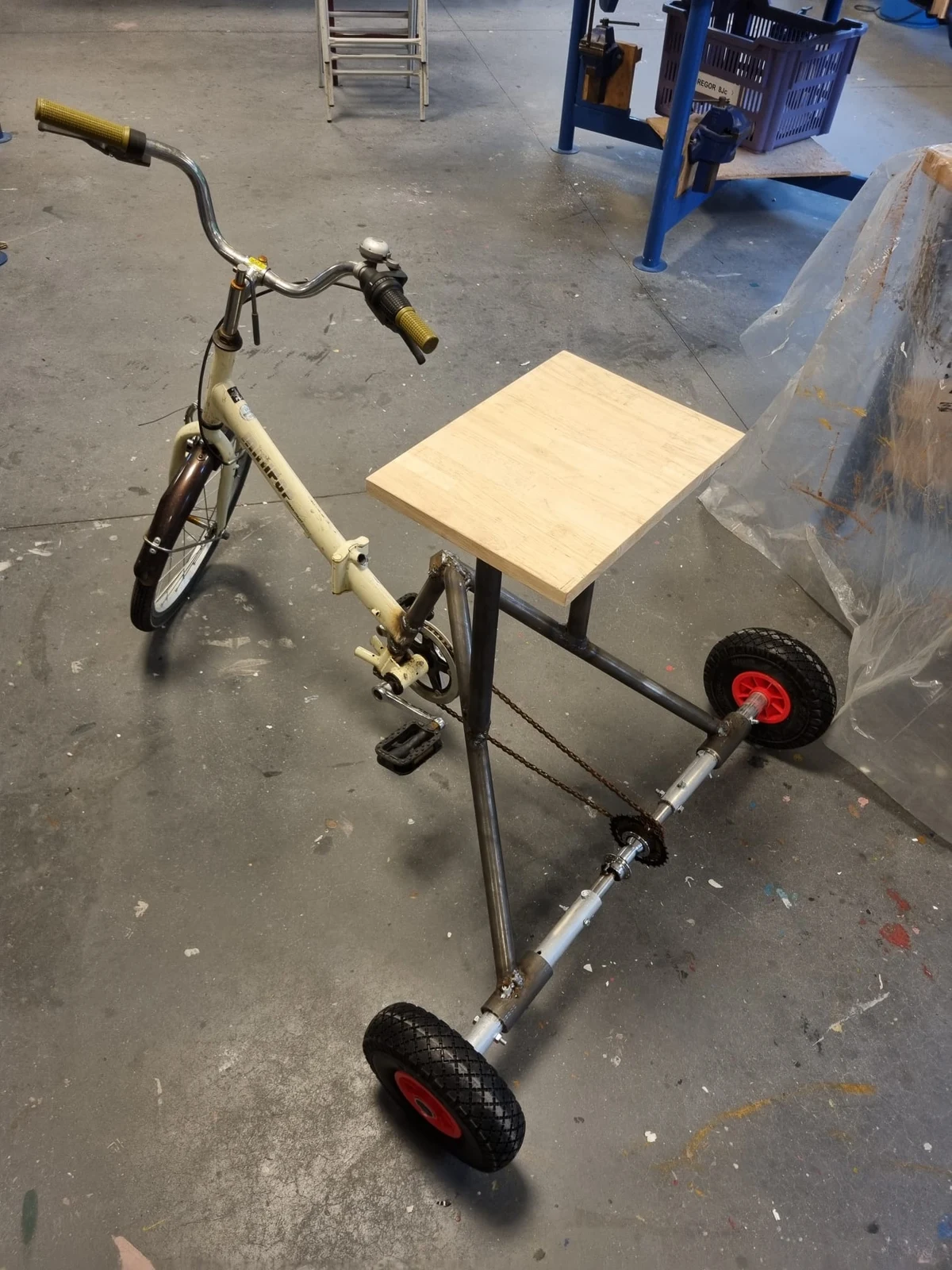 Assembled tricycle in the workshop, with a reused bicycle front, wooden seat, welded rear frame and two small rear wheels.